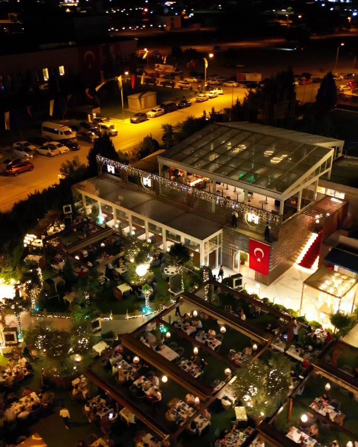 Gece kuş bakışı: bahçedeki dolu masalar ve arkada şehir ışıkları